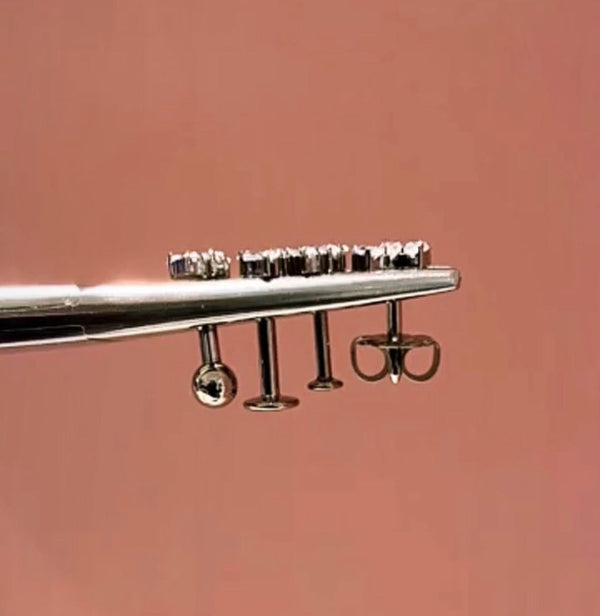 Three piercing jewelry backs compared: ball back, flat disc back, and butterfly earring back — held on forceps against a warm pink background.
